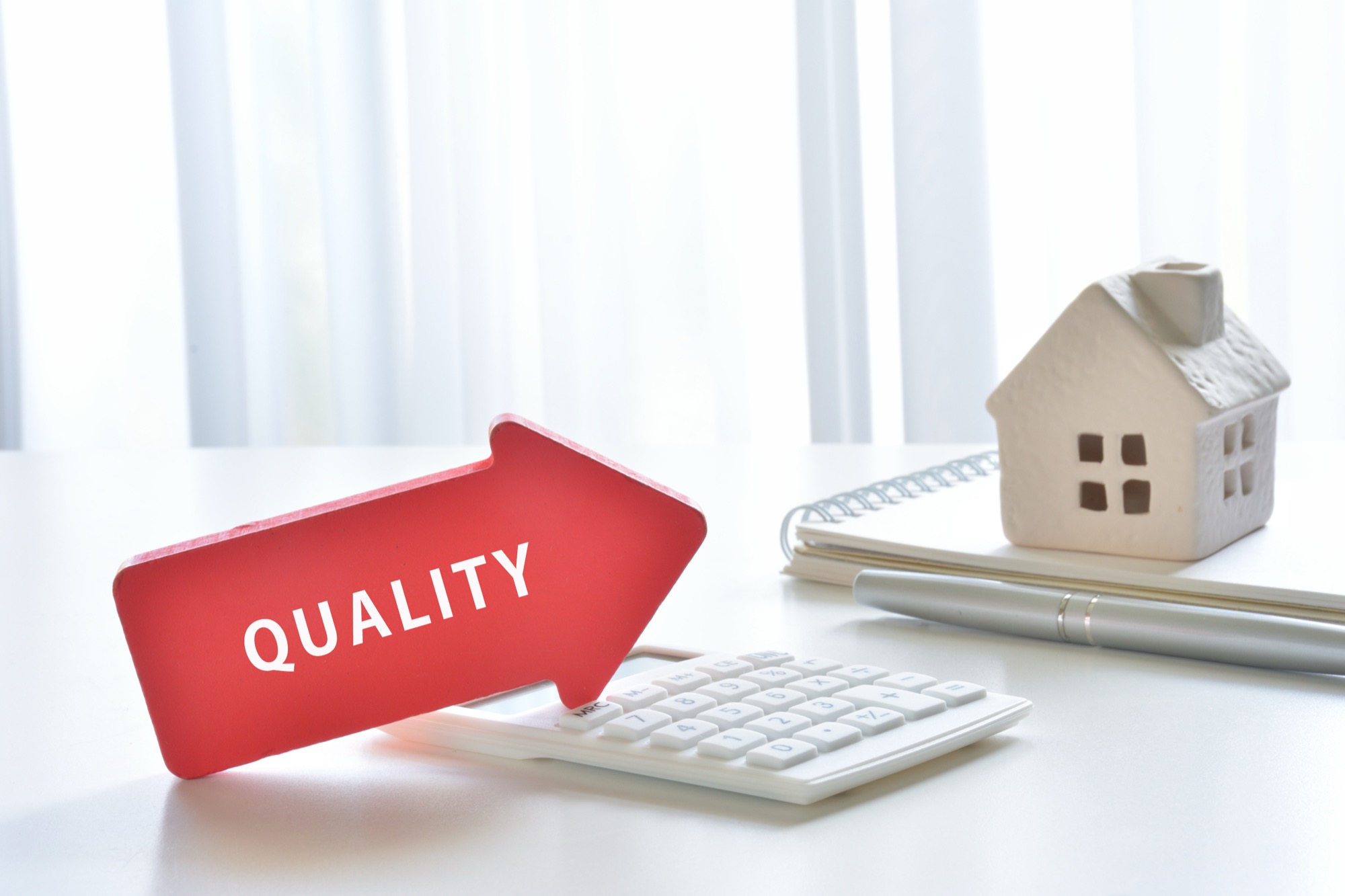 「QUALITY」と書かれた赤い矢印が白い電卓の上に置かれ、奥に白い家型オブジェ、ノート、ペンが並ぶイメージ。