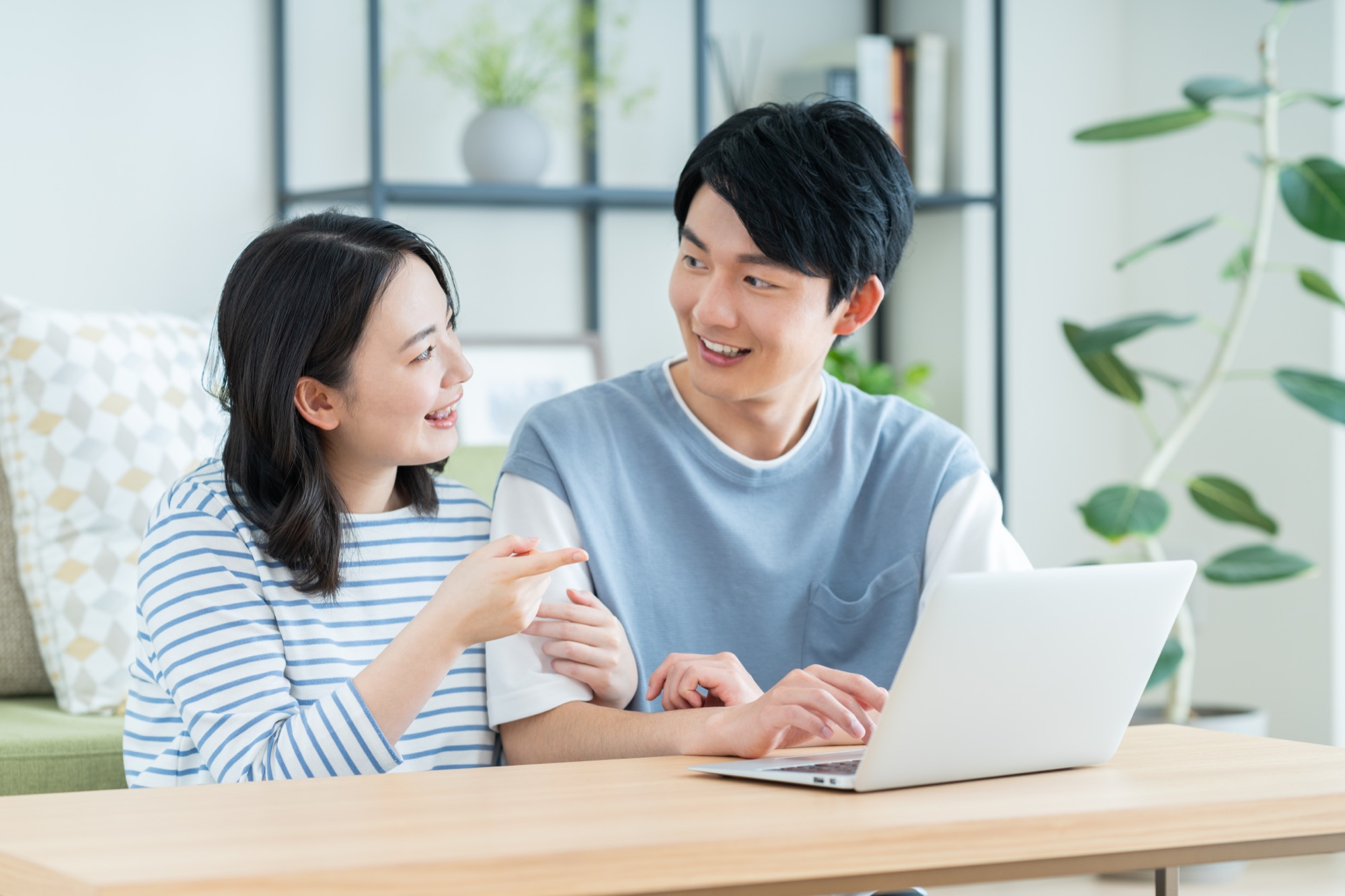 明るいリビングでノートパソコンを囲み、笑顔で会話する若い夫婦。