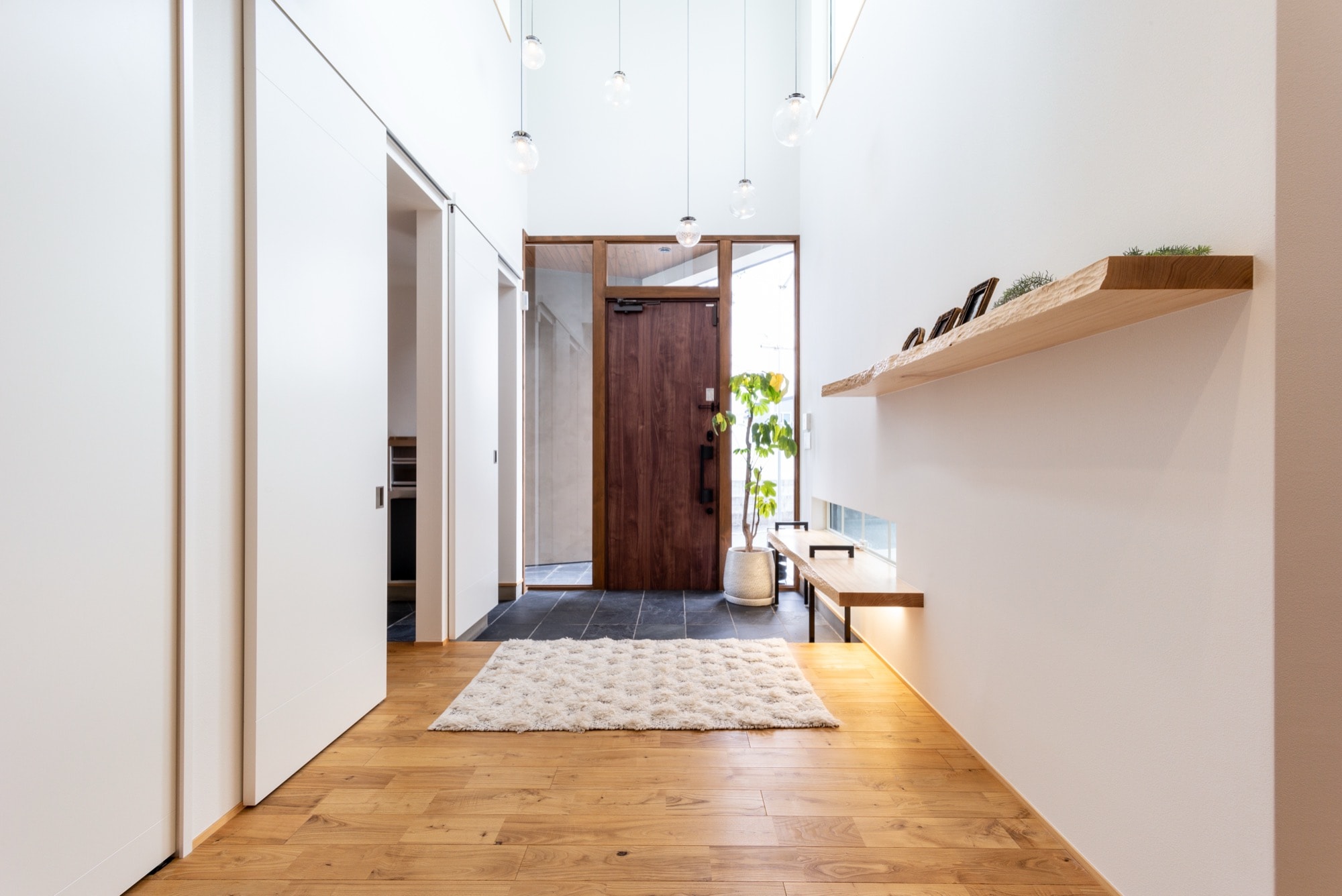 明るく開放的な玄関ホール。高い天井に複数のペンダントライトが吊るされ、温かい木目のフローリングと白い壁が広がる。左手には大容量の白い引き戸収納、右手には間接照明付きの木製飾り棚とベンチが設置され、機能性とデザイン性を両立。玄関土間には黒いタイルと白いラグが敷かれ、モダンな木製玄関ドアと観葉植物が迎える、洗練された住宅の玄関施工事例。