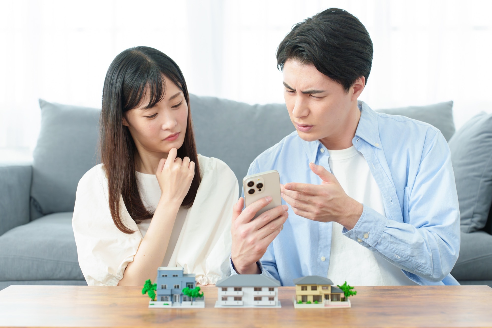 住宅購入を検討する若い夫婦が、スマートフォンで物件情報を見ながら困惑した表情で話し合っている。手前には複数の家とマンションの模型。