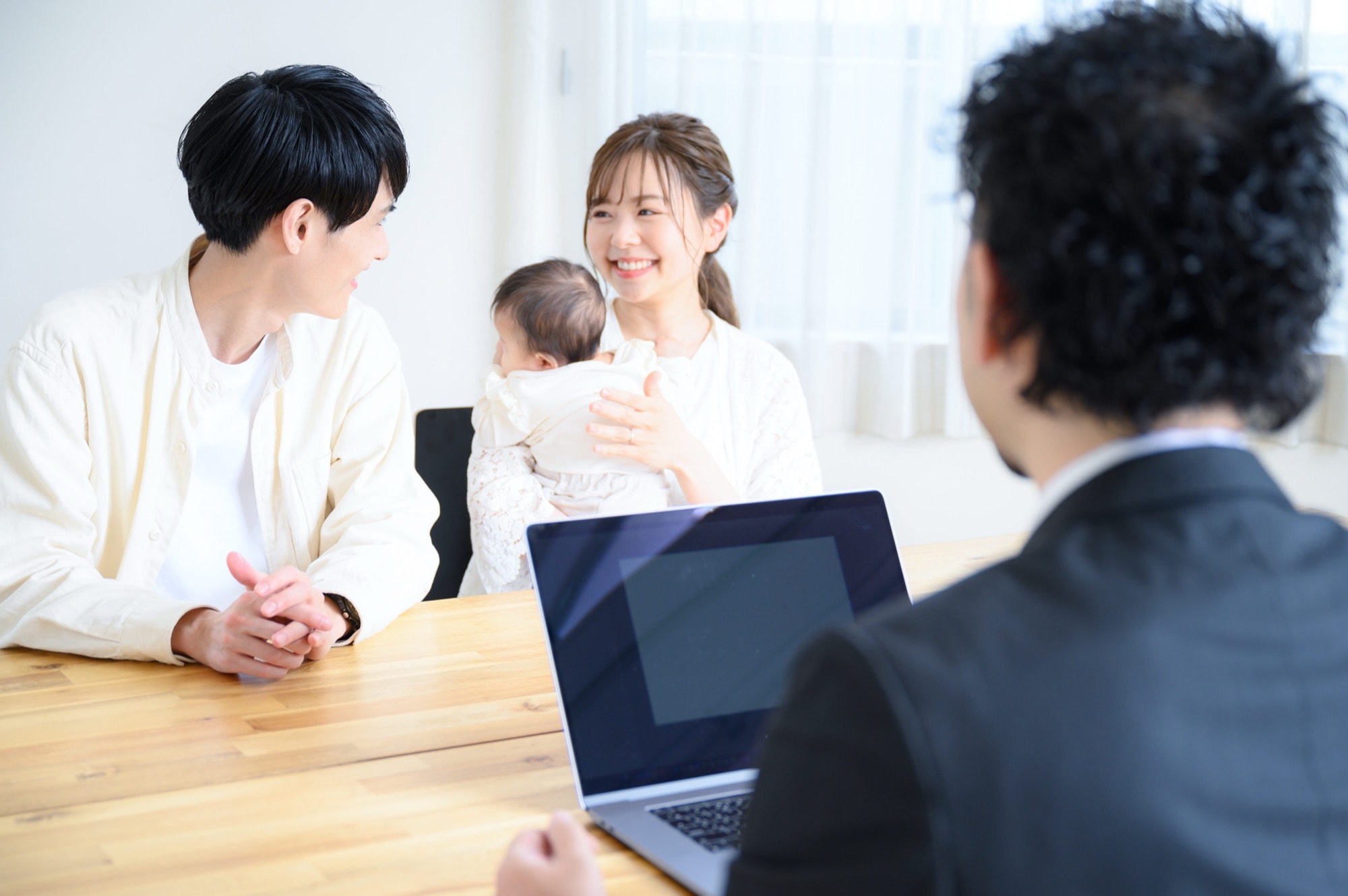 赤ちゃんを抱いた若い夫婦が、専門家とノートパソコンを挟んで笑顔で相談している。明るい室内で、家族のライフプランや未来設計について話し合う和やかな様子。子育て世代の安心感を象徴。