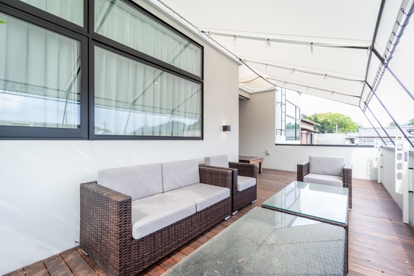 モダンな白い壁と大きな窓に囲まれた広々とした木製デッキテラス。ブラウンのラタン調ソファとアームチェア、ガラス天板のテーブルが配置され、白いオーニングが日差しを遮る快適な屋外リビング空間の施工事例。
