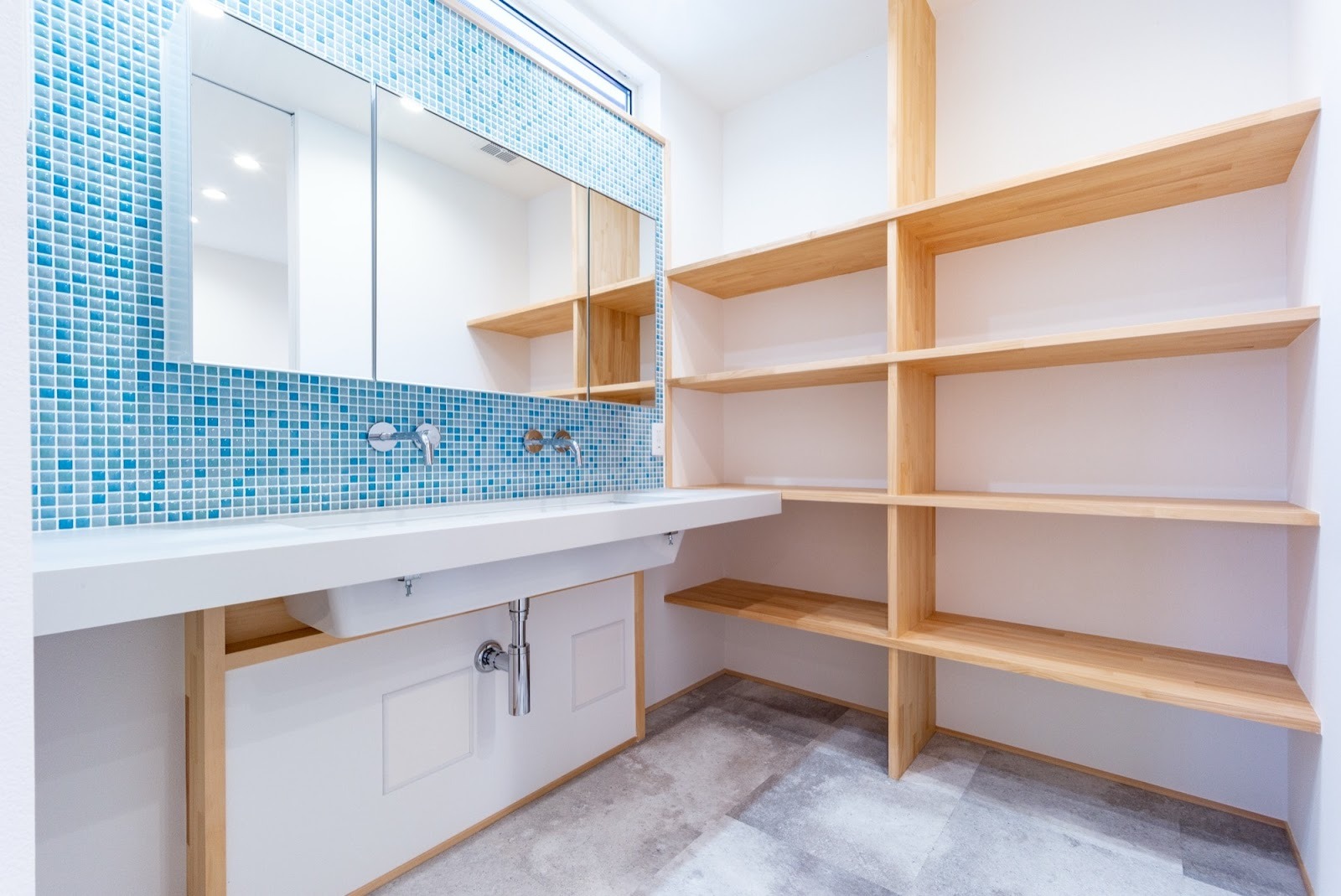 水色のモザイクタイルが壁一面に貼られた、明るく清潔感のある洗面所。2つのシンクを備えた白い洗面台と、天井まで続く木製のオープン収納棚が特徴の、機能性とデザイン性を兼ね備えた洗面空間リフォーム事例。