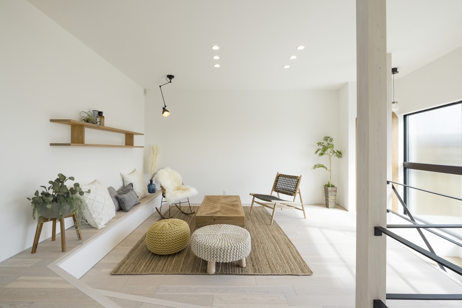 明るい木目フローリングと白い壁で統一された、ナチュラルモダンなリビングルームの施工事例。壁付けベンチソファにはクッションが並び、中央には木製ローテーブル、ジュートラグの上にニットプフやロッキングチェアが配置され、居心地の良い開放的な空間を演出。観葉植物や木製棚の装飾が温かみを添える。