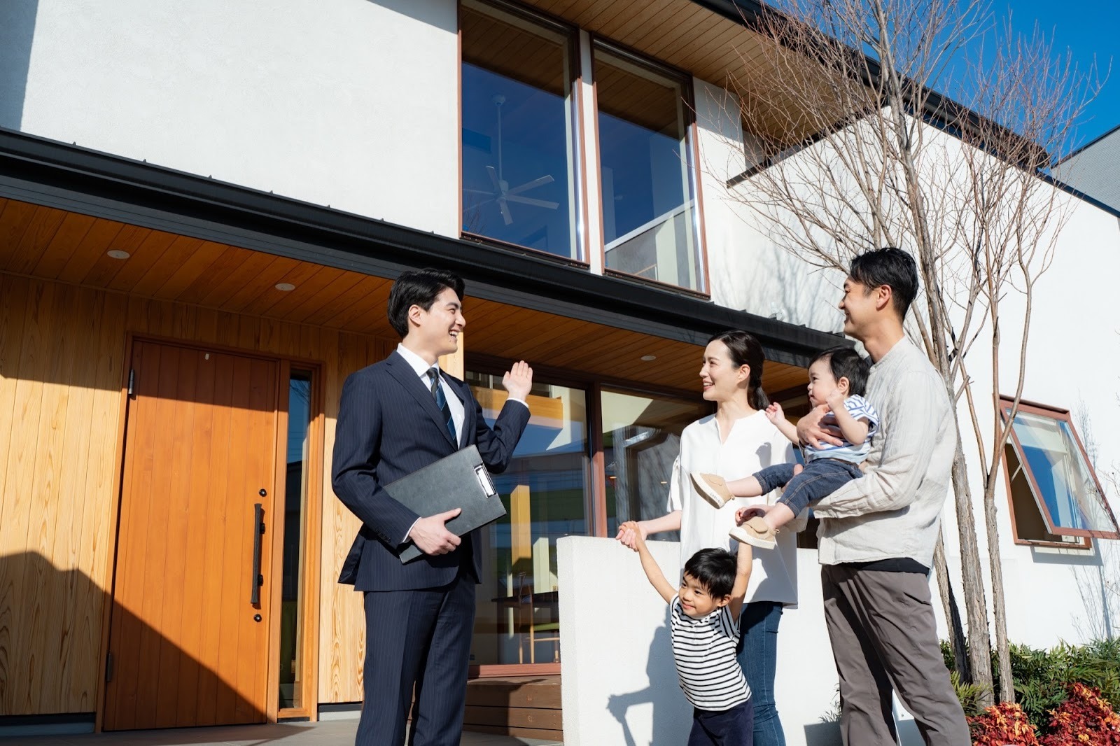 白い外壁と木製ドアのモダンな新築住宅前で、不動産営業担当者が笑顔で若い夫婦と2人の子供に家を案内している。家族は新しいマイホームに期待し、幸せな未来を想像している様子。住宅見学や不動産相談のイメージ。