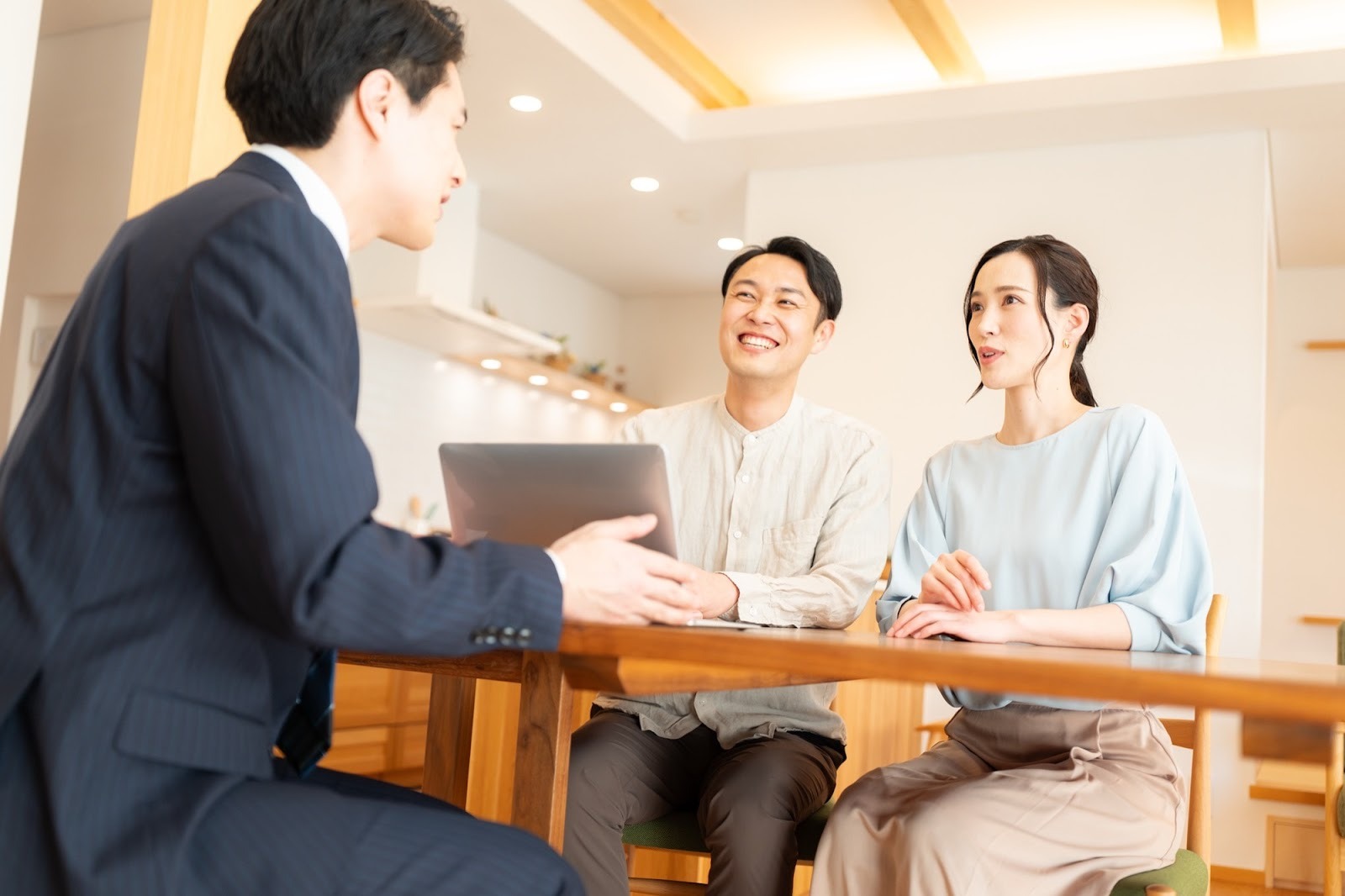 明るいリビングで、ノートパソコンを囲んで笑顔で話す夫婦とスーツ姿の男性。住宅や資産に関する相談やコンサルティングの様子。