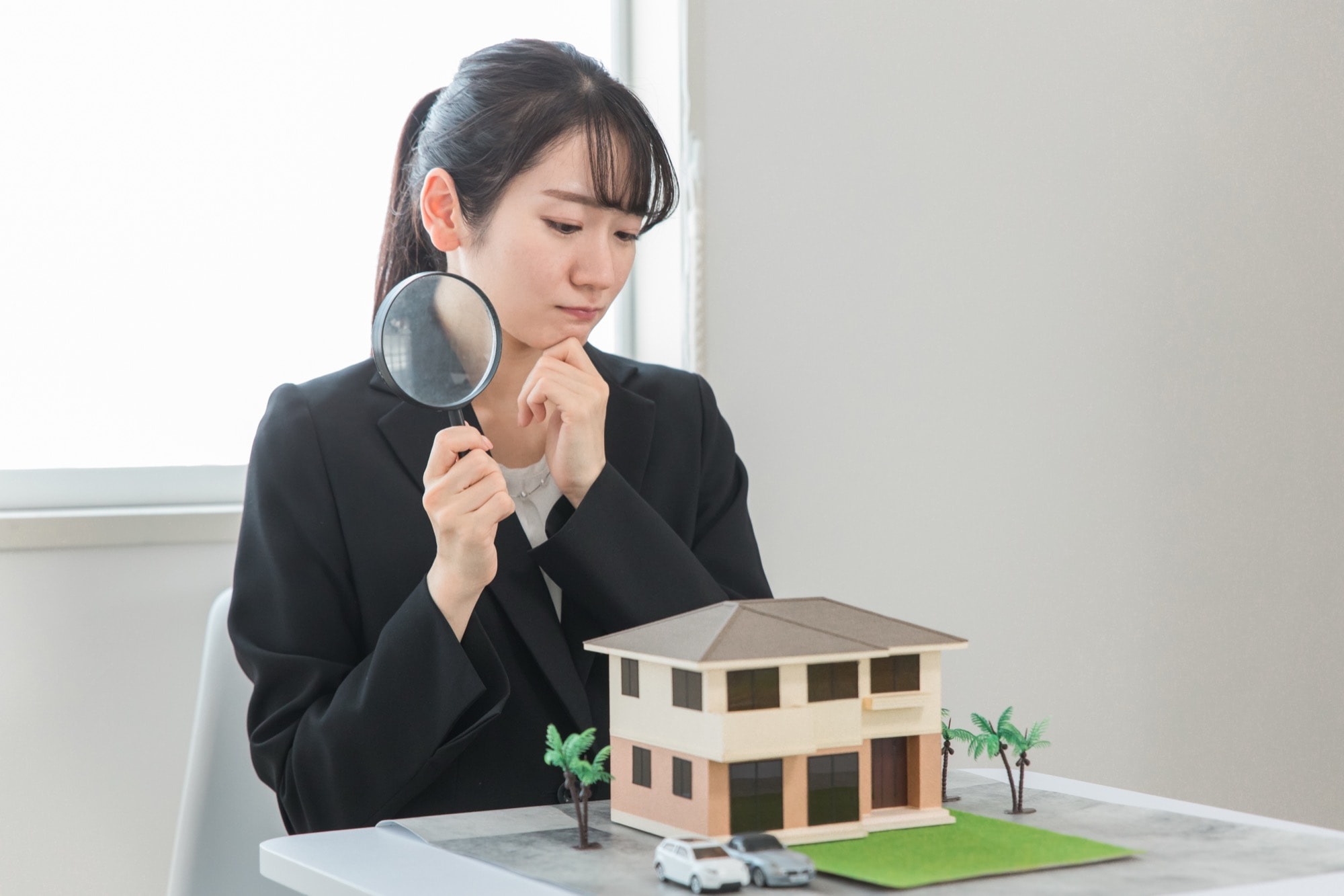 虫眼鏡で家の模型を見ながら悩む女性